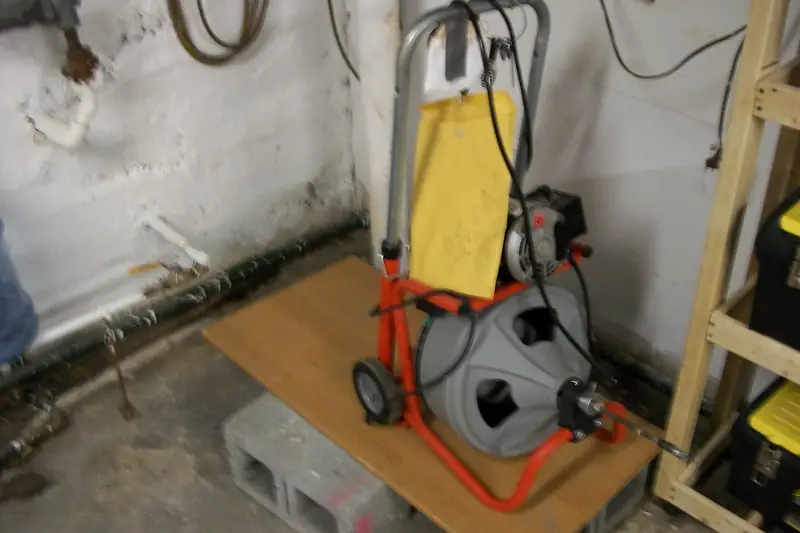 Drum-style drain cleaning machine with its coiled cable, set on the floor next to an open floor drain