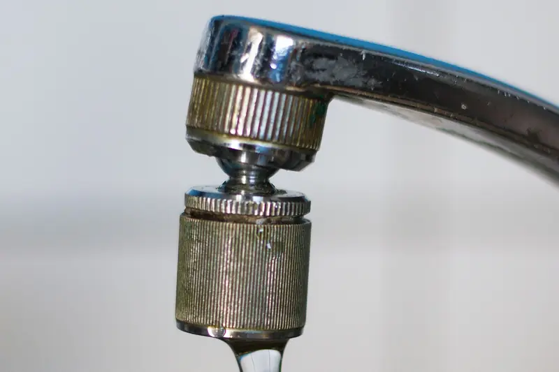 Close-up of a worn chrome faucet spout with a knurled brass aerator and a thin stream of water running from it
