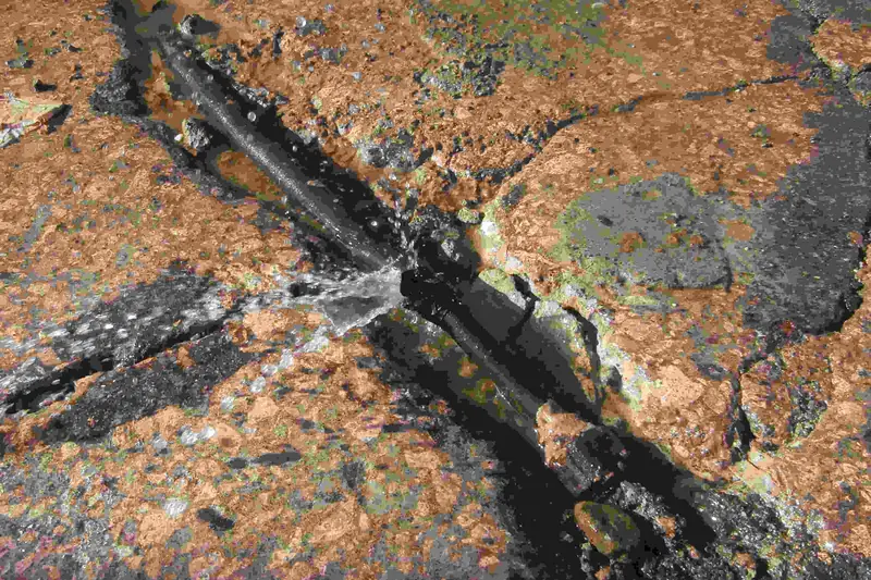 Water spraying from a split in an exposed black pipe lying in a trench of broken pavement and soil