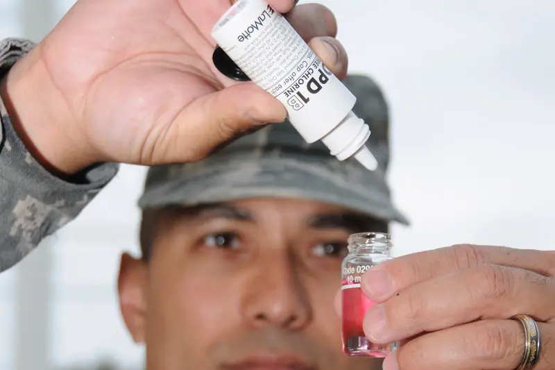 A person holding a small water sample vial tinted pink while squeezing a drop of DPD chlorine test reagent from a white bottle